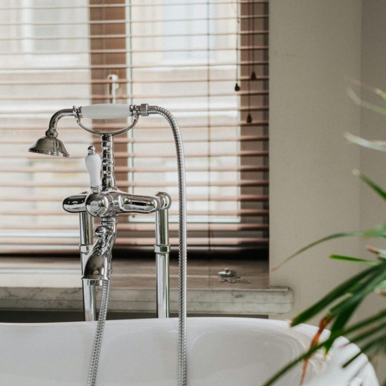 Shower faucet above a white bathtub, with wooden blinds and a plant in the background.