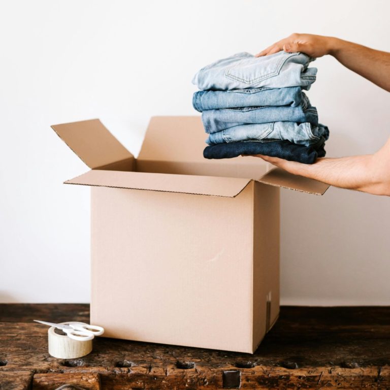 A pair of hands holds neatly folded clothes above an open cardboard box.