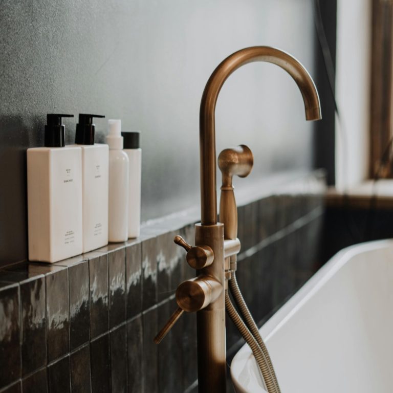 Brass tap and white shampoo bottles beside a modern bathtub against a dark wall.