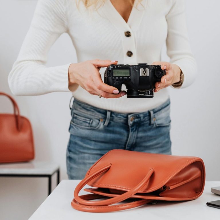 Person holding a camera beside a brown handbag on a table.